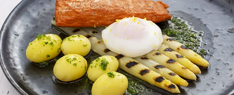 Gegrilde asperges met plantaardige zalm, gepocheerd ei en een kruidige botersaus. Afgewerkt met fijne tuinkruiden en zeste van Amalfi-citroen voor een frisse, culinaire balans.
